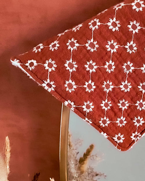 Gros plan sur la lingette en gaze de coton fleurie terracotta, mise en scène avec un fond ocre rouge et quelques tiges de fleurs séchées.