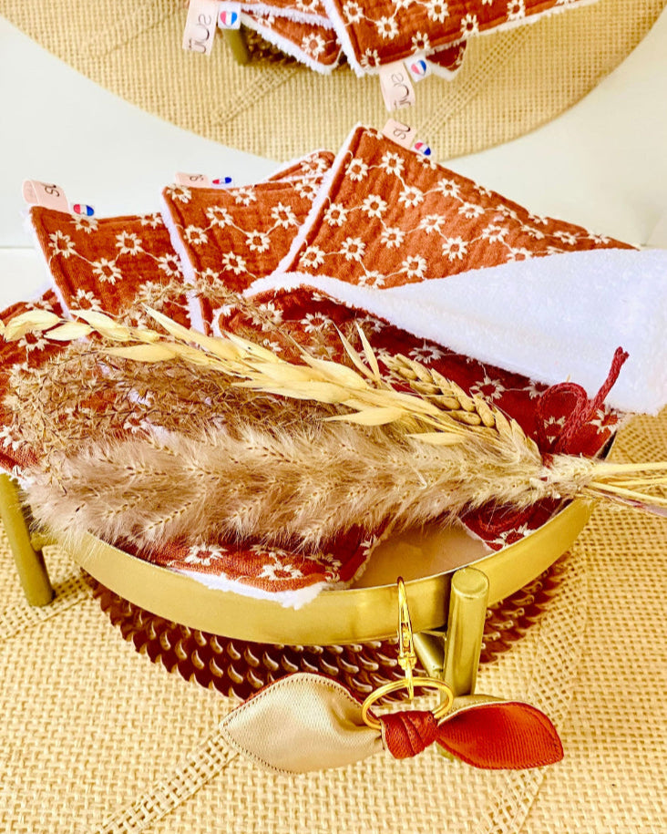 Lingettes démaquillantes en gaze de coton fleurie terracotta exposées sur un plateau doré, accompagnées d’un bouquet de graminées séchées et d’un porte-clé nœud  doré et terracotta